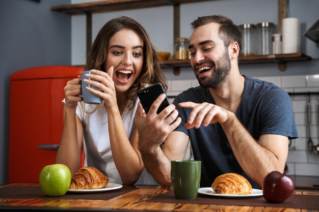 Hermosa pareja alegre desayunando en la cocina, mediante teléfono móvil