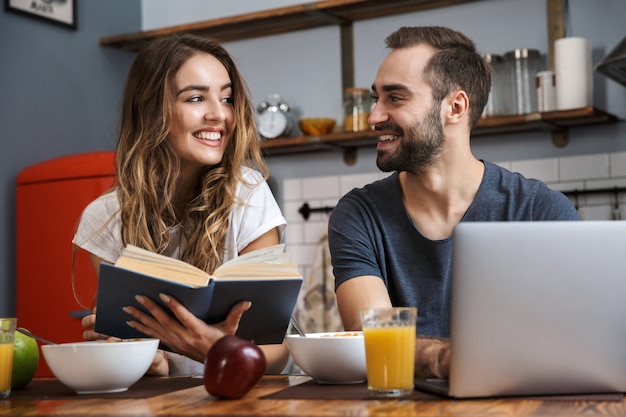 Hermosa pareja alegre desayunando en la cocina, usando la computadora portátil, leyendo un libro