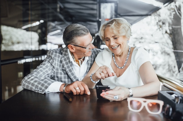 Hermosa pareja de ancianos buscando algo en el teléfono sentado en la terraza de verano en la cafetería moderna y sonriendo sinceramente