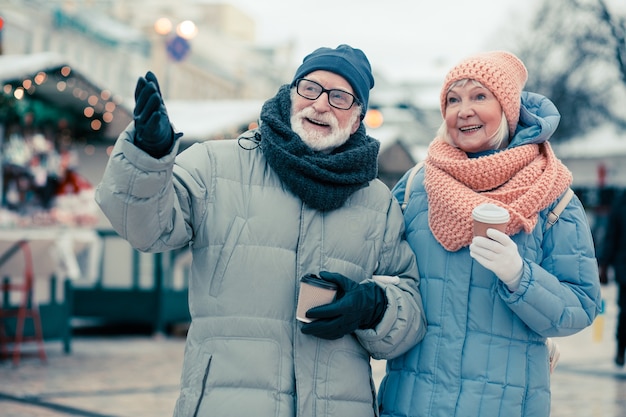 Hermosa pareja de ancianos caminando en día de invierno y disfrutando de un café. Hombre gesticulando con su mano derecha y apuntando a la distancia