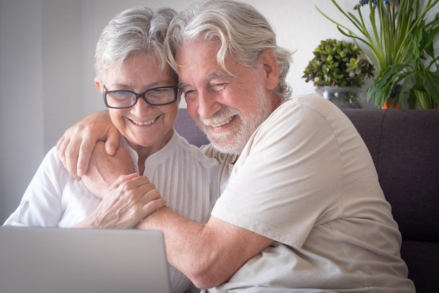 Hermosa pareja de ancianos sentada en casa en un sofá abrazándose mientras navegan en una computadora portátil Personas mayores serenas que disfrutan de la tecnología y navegan socialmente por la red
