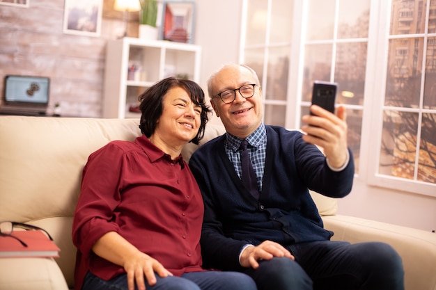 Hermosa pareja de ancianos tomando un selfie mientras está sentado en el sofá de la sala de estar