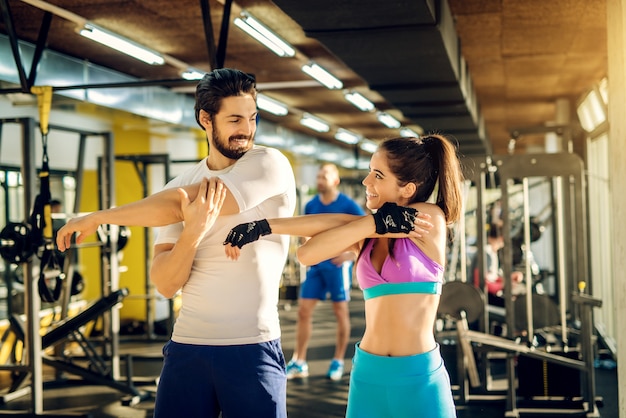 Hermosa pareja atractiva haciendo estiramiento de brazo completo mientras se miran y sonríen en el moderno gimnasio.