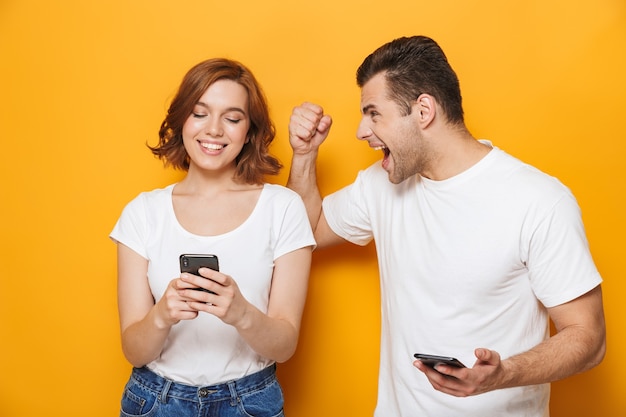 Hermosa pareja emocionada vistiendo camisetas blancas que se encuentran aisladas sobre la pared amarilla, sosteniendo teléfonos móviles, teniendo una pelea