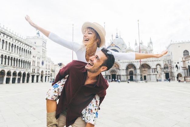 Hermosa pareja de enamorados divirtiéndose abrazando y riendo haciendo caballito en vacaciones en Venecia, Italia, en la Piazza San Marco.