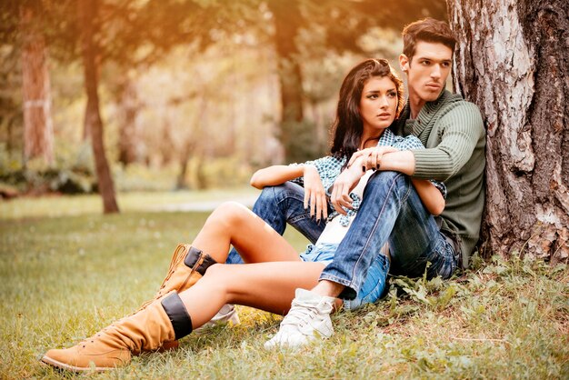 Hermosa pareja encantadora sentada en el césped junto al árbol y disfrutando en un parque soleado en colores otoñales.