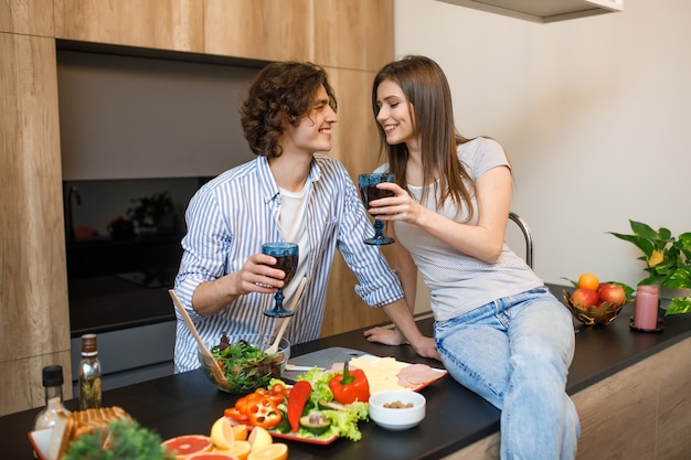 Hermosa pareja está sonriendo mientras bebe vino tinto y cocina juntos en la cocina