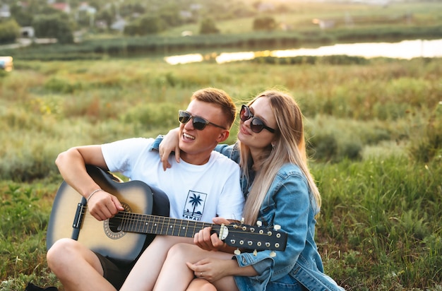 Una hermosa pareja feliz enamorada de gafas de sol tocando la guitarra y regocijándose al atardecer