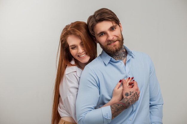 Hermosa pareja feliz de moda en el estudio Feliz hermosa chica pelirroja con sonrisa abrazo guapo brutal hipster hombre con tatuaje y barba en camisa