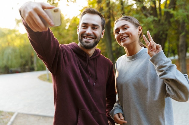 Hermosa pareja feliz en ropa deportiva tomando una foto selfie en el teléfono celular y gesticulando el signo de la paz en el parque verde