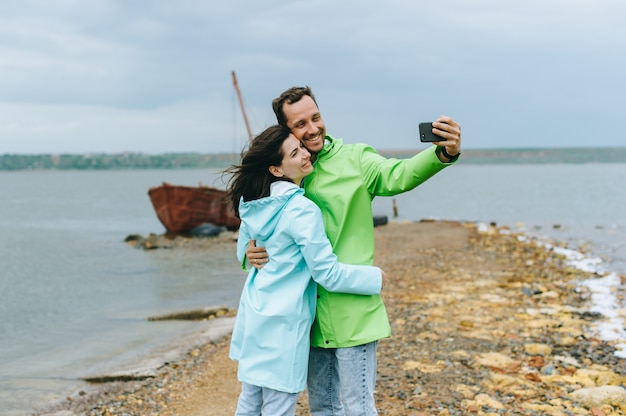 Una hermosa pareja en gabardina de colores hace un selfie cerca del mar.