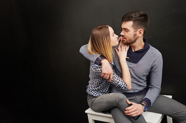 Hermosa pareja de hombre barbudo y mujer rubia sentada contra de fondo negro y mirando el uno al otro