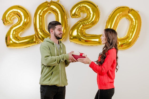 Hermosa pareja hombre y mujer con regalo de Navidad frente a 2020 globos de año nuevo