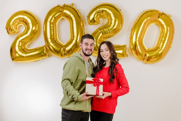 Hermosa pareja hombre y mujer con regalo de Navidad frente a 2020 globos de año nuevo