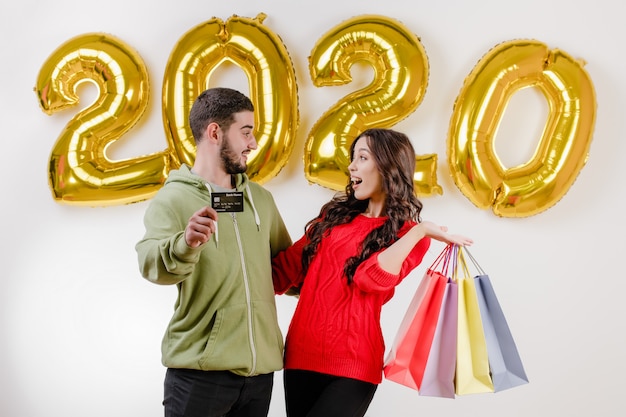 Hermosa pareja hombre y mujer con tarjeta de crédito y coloridas bolsas de compras frente a 2020 globos