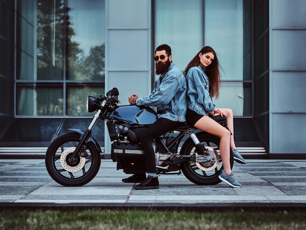 Hermosa pareja interesante está sentada en la bicicleta con chaqueta vaquera cerca del edificio de cristal.