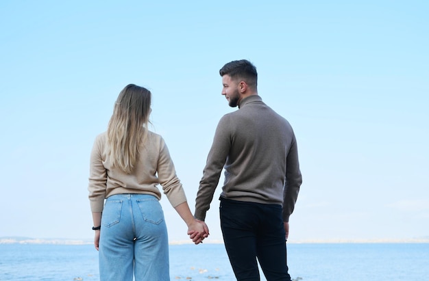 Una hermosa pareja joven se para de espaldas a la cámara y posa frente a un gran lago. El hombre y la mujer se miran con ternura y sonríen.