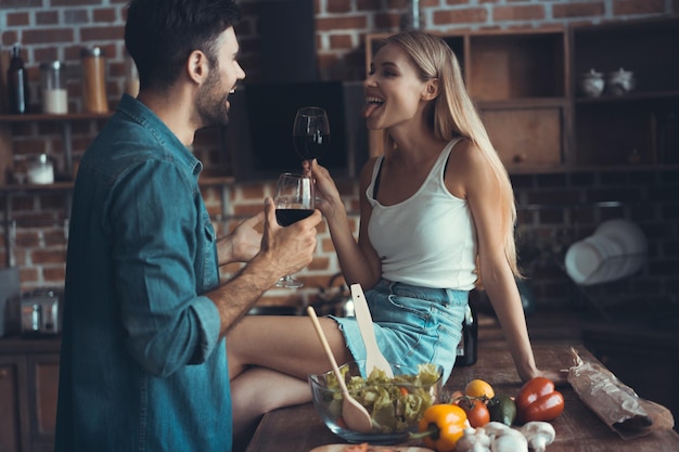 Hermosa pareja joven está bebiendo vino y sonriendo mientras cocina en la cocina en casa