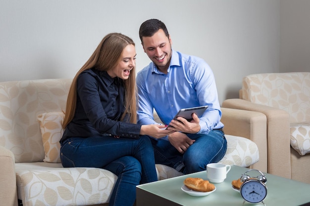 Hermosa pareja joven riéndose, sentada en un sofá, compartiendo fotos u otra información, mostrada en una tableta electrónica y tomando café con croissants