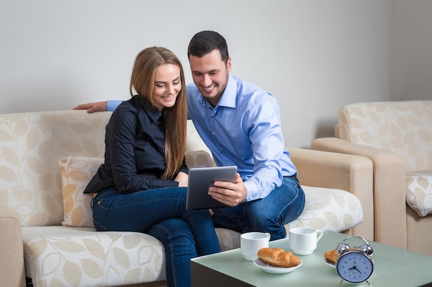 Hermosa pareja joven riéndose, sentada en un sofá, compartiendo fotos u otra información, mostrada en una tableta electrónica y tomando café con croissants