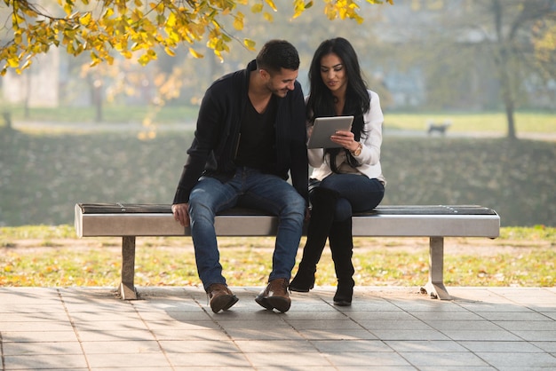 Hermosa pareja joven sentada en un banco en el parque en un hermoso día de otoño que están usando Internet a través de una tableta digital