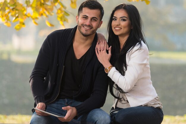 Hermosa pareja joven sentada en un banco en el parque en un hermoso día de otoño que están usando Internet a través de una tableta digital