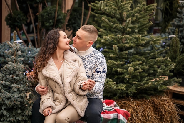 Una hermosa pareja joven sentada cerca de árboles de Navidad con ropa blanca caminando cerca de guirnaldas Feliz hombre y mujer romance Celebración de Navidad diversión amor Suéter blanco año nuevo calle afuera