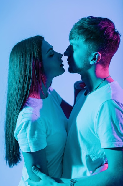 Hermosa pareja joven sexy creativa enamorada en camisetas blancas se besan en un estudio de color sobre un fondo colorido. Chica guapa con chico guapo de moda con luces azules y rosas