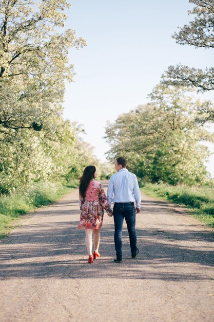 Hermosa pareja joven tomados de la mano y caminando por la carretera bajo el sol entre el campo de primavera y los árboles Familia feliz enamorada relajándose bajo la luz del sol Momentos románticos y alegres Espacio para texto