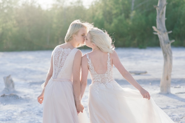 Hermosa pareja de lesbianas caminando sobre la arena a lo largo de la orilla de un río en el día de su boda