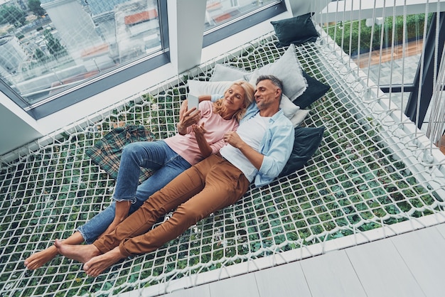 Hermosa pareja madura examinando el teléfono inteligente y sonriendo mientras se relaja en una gran hamaca en casa juntos ...