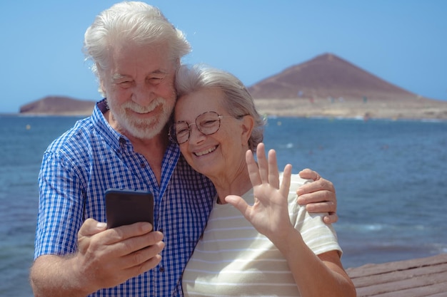 Hermosa pareja mayor caucásica abrazándose mientras hace una videollamada por teléfono móvil frente al mar jubilados mayores disfrutando de vacaciones en el mar en un día soleado y ventoso