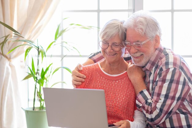 Hermosa pareja mayor caucásica con anteojos navegando juntos en una laptop sentada en casa jubilados ancianos disfrutando de la tecnología y las redes sociales