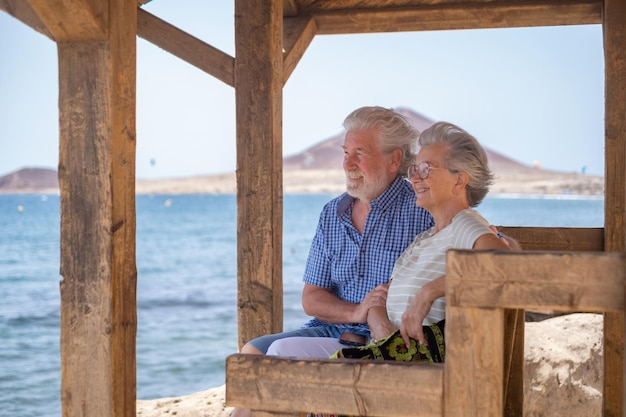 Hermosa pareja mayor caucásica sentada a la sombra de la glorieta frente al mar mirando al horizonte ancianos jubilados disfrutando de vacaciones en el mar en un día ventoso y soleado