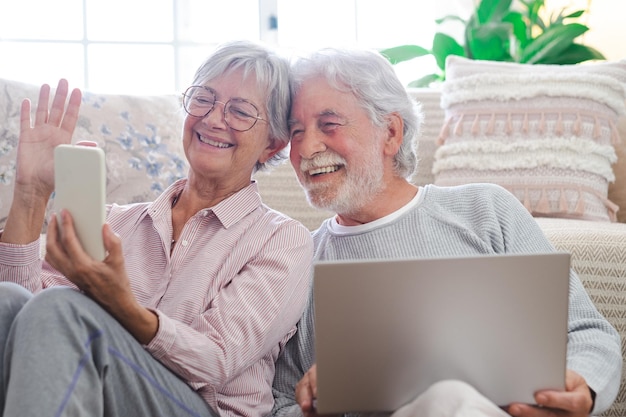 Hermosa pareja mayor caucásica sentada en el suelo en casa usando una computadora portátil y un teléfono móvil modernos jubilados ancianos usando dispositivos tecnológicos