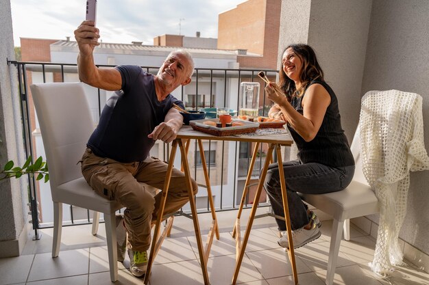 Foto una hermosa pareja de mediana edad comiendo comida china y tomando una selfie en una mesa en un balcón de la ciudad