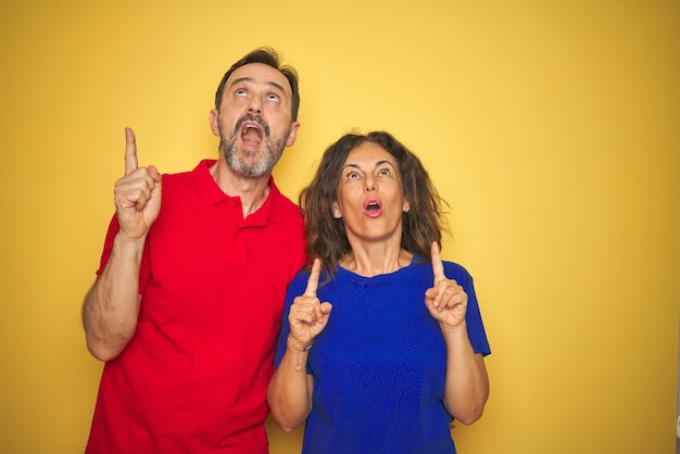 Hermosa pareja de mediana edad juntos de pie sobre un fondo amarillo aislado asombrado y sorprendido mirando hacia arriba y señalando con los dedos y los brazos levantados