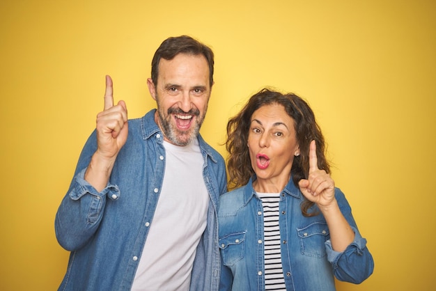 Hermosa pareja de mediana edad juntos usando camisa de mezclilla sobre fondo amarillo aislado señalando con el dedo hacia arriba con una idea exitosa Exitosa y feliz Número uno