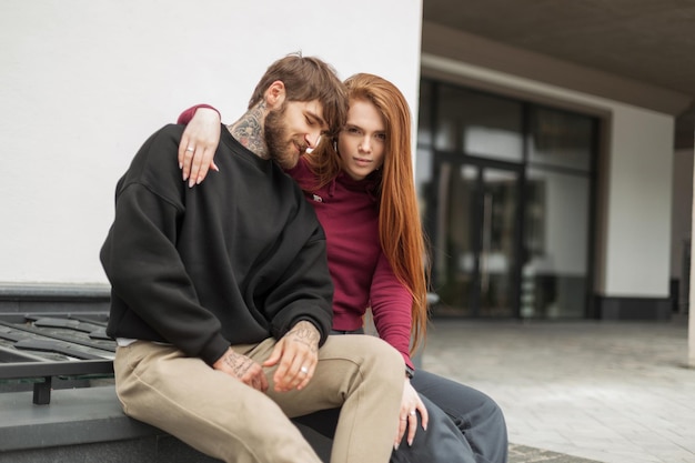 Foto hermosa pareja de moda brutal barbudo hipster hombre guapo y hermosa chica de pelo rojo lindo en ropa de moda están sentados en la calle