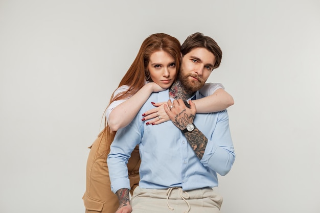 Hermosa pareja de moda, un brutal hombre hipster con barba y tatuajes y una hermosa chica pelirroja con una camisa sobre un fondo blanco. Un chico guapo se sienta en una silla y una mujer lo abraza.