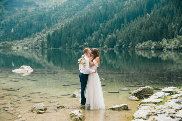 Hermosa pareja moderna cerca de un lago en las montañas hacen fotos de boda