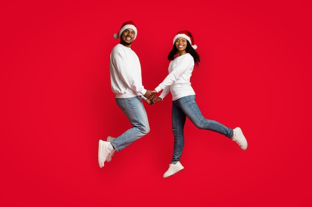 Hermosa pareja negra con sombreros de Santa cogidos de la mano y saltando