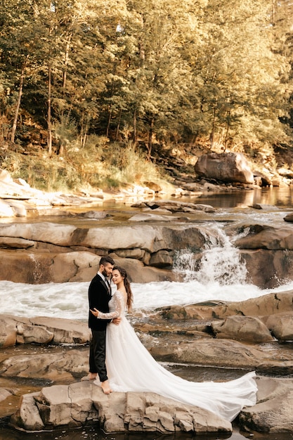 Hermosa pareja de novios se abrazan tiernamente en el fondo de un río de montaña, vestido largo y blanco de la novia.