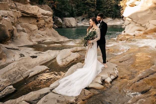 Hermosa pareja de novios se abrazan tiernamente en el fondo de un río de montaña, vestido largo y blanco de la novia.