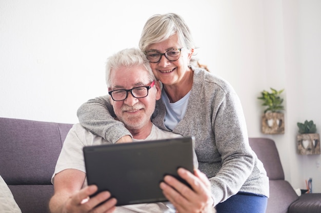 Hermosa pareja de personas mayores o personas maduras en casa abrazados mirando la misma tableta o computadora portátil: jubila a las personas mayores con gafas usando la nueva tecnología