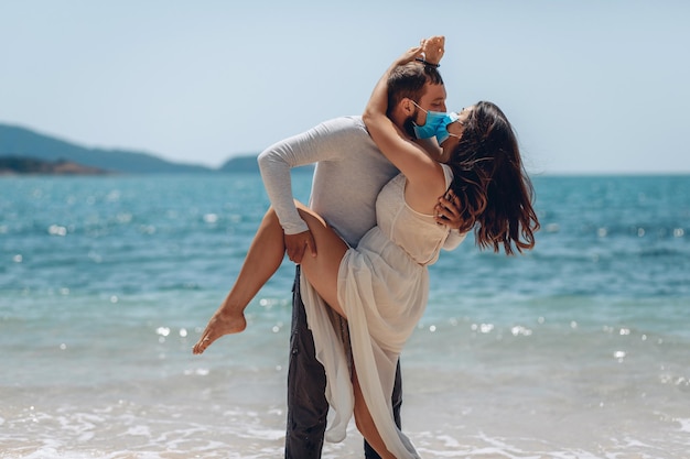 Hermosa pareja romántica besándose juguetonamente en la playa con máscaras médicas que los protegen de un brote de infección por coronavirus. Hombre sujetando la pierna de su novia y besándola