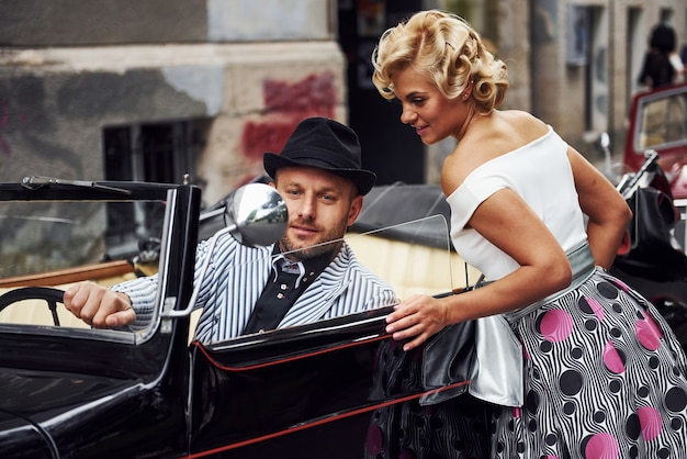 Foto hermosa pareja en ropa antigua está en la ciudad con coche retro.