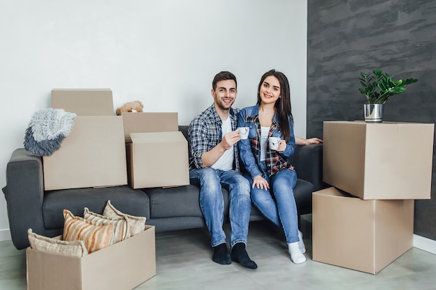 Hermosa pareja en ropa casual está discutiendo el plan de su nueva casa y sonriendo mientras está acostado en el sofá cerca de las cajas para moverse. El hombre esta tomando cafe