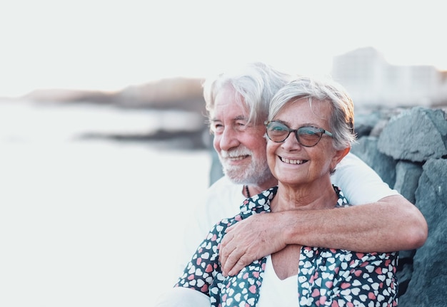 Hermosa pareja senior abrazándose sonriendo al aire libre en el mar mirando a otro lado. Pareja caucásica de pelo blanco disfrutando de la jubilación y la luz del atardecer