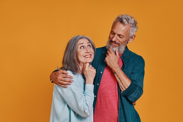 Hermosa pareja senior abrazándose y sonriendo mientras están de pie juntos contra el fondo naranja